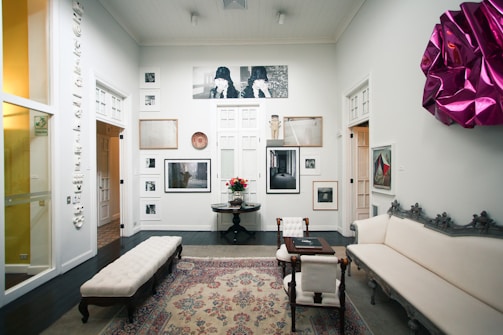A well-decorated room featuring a variety of artwork on the walls, including photographs and framed art pieces. There is an elegant rug on the dark wooden floor, and the space is furnished with a white upholstered couch, a coffee table, and chairs. A vibrant magenta wall decoration adds a pop of color to the otherwise neutral color scheme. Soft lighting enhances the calm and sophisticated ambiance.