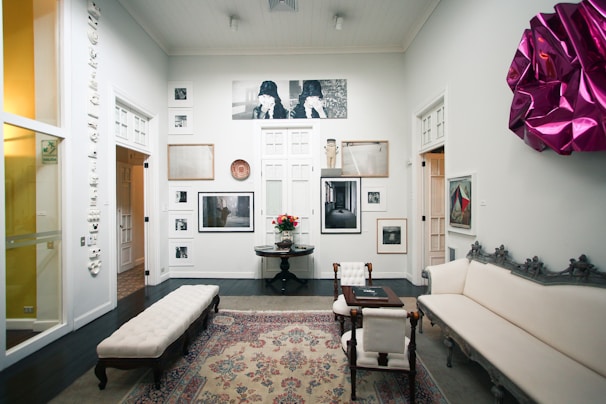 A well-decorated room featuring a variety of artwork on the walls, including photographs and framed art pieces. There is an elegant rug on the dark wooden floor, and the space is furnished with a white upholstered couch, a coffee table, and chairs. A vibrant magenta wall decoration adds a pop of color to the otherwise neutral color scheme. Soft lighting enhances the calm and sophisticated ambiance.