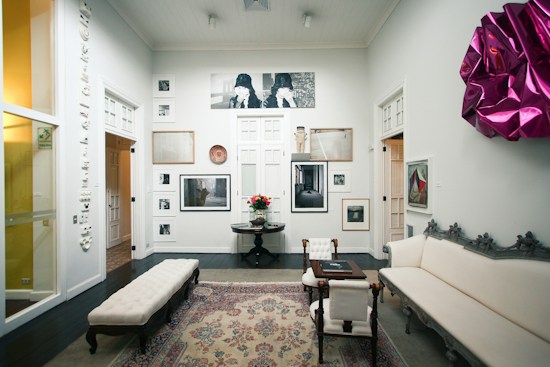 A well-decorated room featuring a variety of artwork on the walls, including photographs and framed art pieces. There is an elegant rug on the dark wooden floor, and the space is furnished with a white upholstered couch, a coffee table, and chairs. A vibrant magenta wall decoration adds a pop of color to the otherwise neutral color scheme. Soft lighting enhances the calm and sophisticated ambiance.
