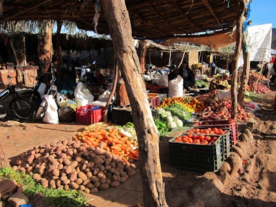 A bustling outdoor market stall with various vegetables, including potatoes, carrots, tomatoes, and onions, arranged in piles and crates. The market is sheltered by a rustic wooden canopy, and motorbikes can be seen in the background. People are busy and engaged, indicating daily market activity.