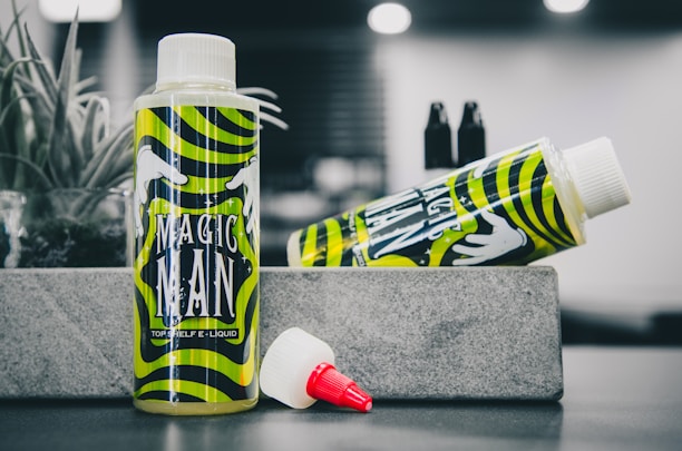 A bottle of e-liquid labeled 'Magic Man' is prominently displayed against a blurred background. The design features vibrant green and yellow swirling patterns with white hand illustrations. Next to the upright bottle, another similar bottle lies on its side. In the foreground, there is a red-tipped, white nozzle cap. An air plant sits to the side on the stone-like surface.