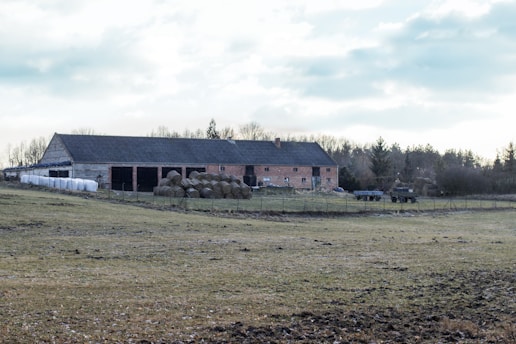 A rural landscape features a large barn and adjacent storage building, both constructed from brick and stone. In front of the buildings, there are several cylindrical bales of hay neatly arranged. Open fields of grass and dirt extend in the foreground, leading to a slightly overcast sky. The scene has a tranquil, pastoral setting typical of a countryside farm.