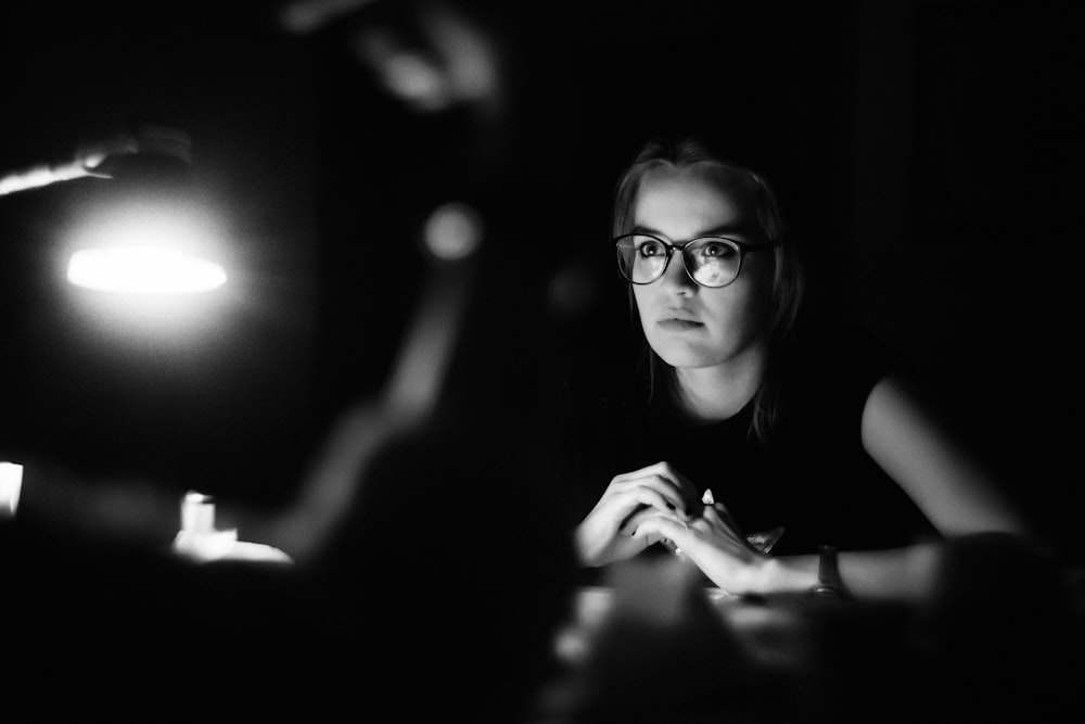 grayscale photo of a woman wearing eyeglasses sitting in front of table