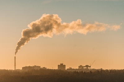 A factory chimney releases a thick plume of smoke into the sky, silhouetted against a hazy, orange-toned sunset or sunrise. The smoke trails diagonally across the sky, suggesting pollution. In the background, urban buildings and construction equipment, such as cranes, are visible.