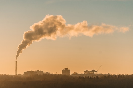 A factory chimney releases a thick plume of smoke into the sky, silhouetted against a hazy, orange-toned sunset or sunrise. The smoke trails diagonally across the sky, suggesting pollution. In the background, urban buildings and construction equipment, such as cranes, are visible.