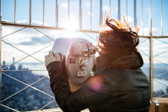 A person with long hair blowing in the wind stands on a high vantage point holding a coin-operated binocular viewer. The background reveals a cityscape under a clear sky with bright sunlight creating a lens flare effect.