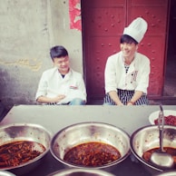 Two young men wearing white chef jackets are seated outside in front of a rustic wall and door. The one on the right is wearing a chef hat and is smiling, while the other looks on with a slight smile. In front of them are several large metal bowls filled with a spicy red dish, featuring visible chili peppers and oil.