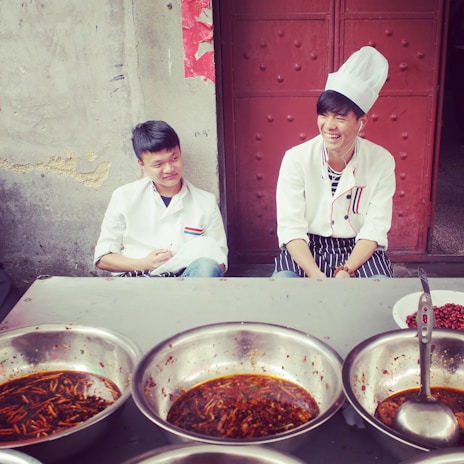 Two young men wearing white chef jackets are seated outside in front of a rustic wall and door. The one on the right is wearing a chef hat and is smiling, while the other looks on with a slight smile. In front of them are several large metal bowls filled with a spicy red dish, featuring visible chili peppers and oil.