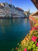 A scenic riverside view featuring historic European buildings with ornate facades, a Swiss flag on a rooftop, and a spire in the background. The foreground includes a vibrant display of red and pink flowers along a wooden walkway. A blue sky and calm, reflective water add to the tranquil setting.