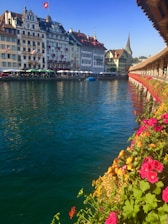 A scenic riverside view featuring historic European buildings with ornate facades, a Swiss flag on a rooftop, and a spire in the background. The foreground includes a vibrant display of red and pink flowers along a wooden walkway. A blue sky and calm, reflective water add to the tranquil setting.