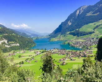 A picturesque landscape features a turquoise lake surrounded by lush green hills. A cluster of small houses is nestled in the valley, and towering mountains frame the serene scene under a clear blue sky.