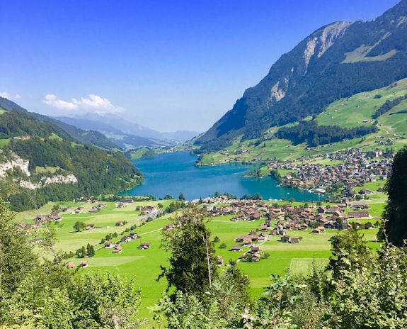 A picturesque landscape features a turquoise lake surrounded by lush green hills. A cluster of small houses is nestled in the valley, and towering mountains frame the serene scene under a clear blue sky.