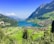 A picturesque landscape features a turquoise lake surrounded by lush green hills. A cluster of small houses is nestled in the valley, and towering mountains frame the serene scene under a clear blue sky.