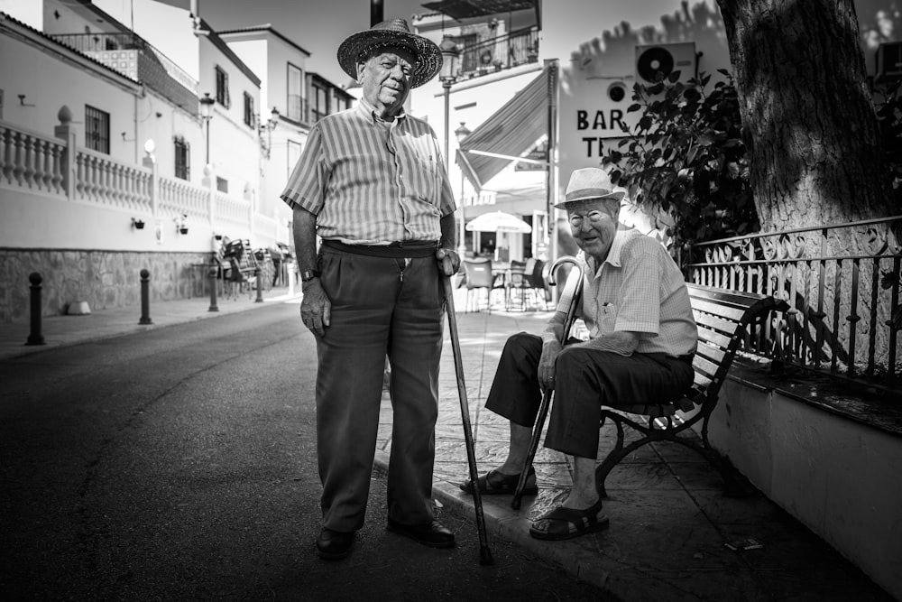 man standing beside a man sitting on cast iron bench near buildings