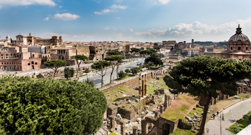 An expansive view of ancient Roman ruins with a mixture of dark green trees and historic buildings. The skyline reveals classic and Renaissance architecture amidst a clear sky. A roadway runs through the center, lined with more of the city's historical structures.