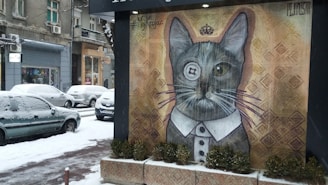 A street art mural depicting a cat with a monocle and a suit collar, giving it a sophisticated appearance. The mural is set against a textured background and is located in an urban area with snowy weather. Several cars are parked along the snowy road, and the buildings in the background have a classical architectural style.