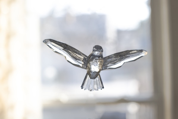 A crystal sculpture of a bird with outstretched wings, set against a softly blurred background. The intricate facets of the crystal play with the light, creating reflections and highlighting the bird's detailed design.