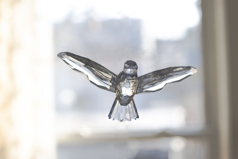 A crystal sculpture of a bird with outstretched wings, set against a softly blurred background. The intricate facets of the crystal play with the light, creating reflections and highlighting the bird's detailed design.