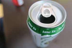 A close-up of an opened can of amber ale with a green and white design, featuring text indicating the type of beverage. A blurred object in a red color is visible in the background.