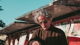 A person with gray hair and sunglasses is seen under a metal awning. They are wearing a brown knitted sweater and appear to be engaged in an activity, possibly selling or inspecting items. Various artworks or prints are clipped and displayed in the background, adding vibrant colors to the scene.