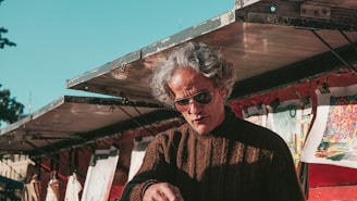 A person with gray hair and sunglasses is seen under a metal awning. They are wearing a brown knitted sweater and appear to be engaged in an activity, possibly selling or inspecting items. Various artworks or prints are clipped and displayed in the background, adding vibrant colors to the scene.
