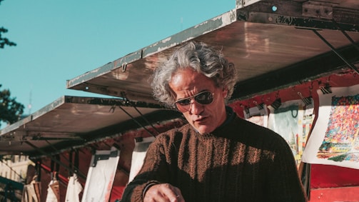 A person with gray hair and sunglasses is seen under a metal awning. They are wearing a brown knitted sweater and appear to be engaged in an activity, possibly selling or inspecting items. Various artworks or prints are clipped and displayed in the background, adding vibrant colors to the scene.