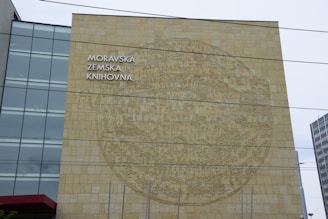 A large building façade with a detailed circular mural etched into a stone wall. The words 'MORAVSKÁ ZEMSKÁ KNIHOVNA' are featured prominently in block letters. To the left, there is a modern section of the building with glass windows.