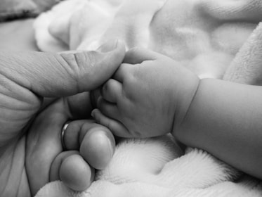 An adult hand gently holds a baby's small hand, conveying a sense of tenderness and connection. The background consists of soft fabric, enhancing the intimate moment between the two.