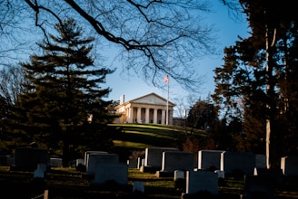 A stately building with classical columns sits atop a grassy hill, flanked by tall trees. A flagpole in front displays the American flag at half-mast. The foreground features numerous headstones, suggesting a cemetery setting, with leafless branches framing the scene.