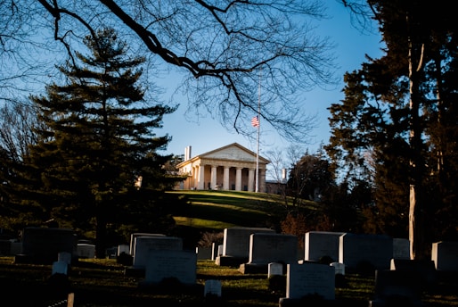 A stately building with classical columns sits atop a grassy hill, flanked by tall trees. A flagpole in front displays the American flag at half-mast. The foreground features numerous headstones, suggesting a cemetery setting, with leafless branches framing the scene.