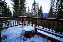 A wooden balcony overlooks a snowy forest landscape with tall evergreen trees. A small round table and a bench, both dusted with snow, are positioned on the balcony, which has a simple wooden railing. In the background, snow-covered mountains are visible under a clear sky.