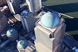 An aerial view of skyscrapers with distinct turquoise domes on the rooftops. The tall buildings are positioned near a waterfront, which can be seen in the background. Streets with visible yellow taxis line the ground below.