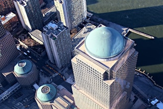 An aerial view of skyscrapers with distinct turquoise domes on the rooftops. The tall buildings are positioned near a waterfront, which can be seen in the background. Streets with visible yellow taxis line the ground below.