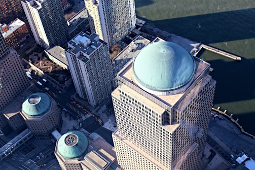 An aerial view of skyscrapers with distinct turquoise domes on the rooftops. The tall buildings are positioned near a waterfront, which can be seen in the background. Streets with visible yellow taxis line the ground below.