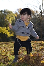 A young child is standing on a bed of fallen leaves in an outdoor setting, holding a stick in one hand. The child is wearing a grey hoodie with wooden toggles, a yellow shirt, black pants, and grey boots. The background features trees with autumn foliage and a clear sky.
