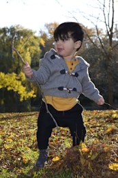 A young child is standing on a bed of fallen leaves in an outdoor setting, holding a stick in one hand. The child is wearing a grey hoodie with wooden toggles, a yellow shirt, black pants, and grey boots. The background features trees with autumn foliage and a clear sky.