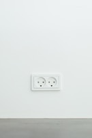 A white electrical wall socket with two outlets is mounted on a plain white wall with a grey baseboard at the bottom.