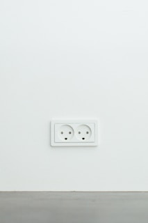 A white electrical wall socket with two outlets is mounted on a plain white wall with a grey baseboard at the bottom.