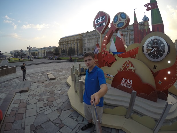 A young man in a blue shirt is taking a selfie with a large decorative installation. The installation has clocks showing a countdown and the logo for the 2018 FIFA World Cup Russia. The background displays a public square with historical buildings and people walking.