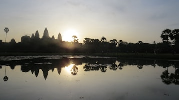 A serene scene captures a silhouette of ancient temple towers with the sun setting or rising behind them, casting a warm glow. In the foreground, a calm body of water reflects the structure and sky, while palm trees are visible on both sides. The image exudes tranquility and historical grandeur.
