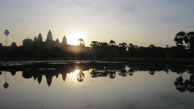 A serene scene captures a silhouette of ancient temple towers with the sun setting or rising behind them, casting a warm glow. In the foreground, a calm body of water reflects the structure and sky, while palm trees are visible on both sides. The image exudes tranquility and historical grandeur.