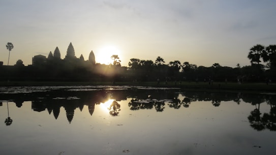 A serene scene captures a silhouette of ancient temple towers with the sun setting or rising behind them, casting a warm glow. In the foreground, a calm body of water reflects the structure and sky, while palm trees are visible on both sides. The image exudes tranquility and historical grandeur.