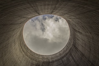 An upward view from inside a cooling tower shows a circular opening towards the cloudy sky, revealing the towering concrete structure's textured, ribbed surface.