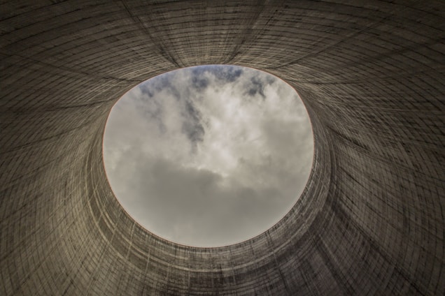 An upward view from inside a cooling tower shows a circular opening towards the cloudy sky, revealing the towering concrete structure's textured, ribbed surface.
