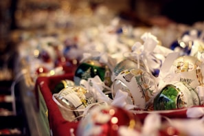 A collection of Christmas ornaments displayed in a basket, featuring intricate designs and festive illustrations. The decorations are adorned with ribbons and showcase a mix of colors and patterns, creating a warm, holiday atmosphere.