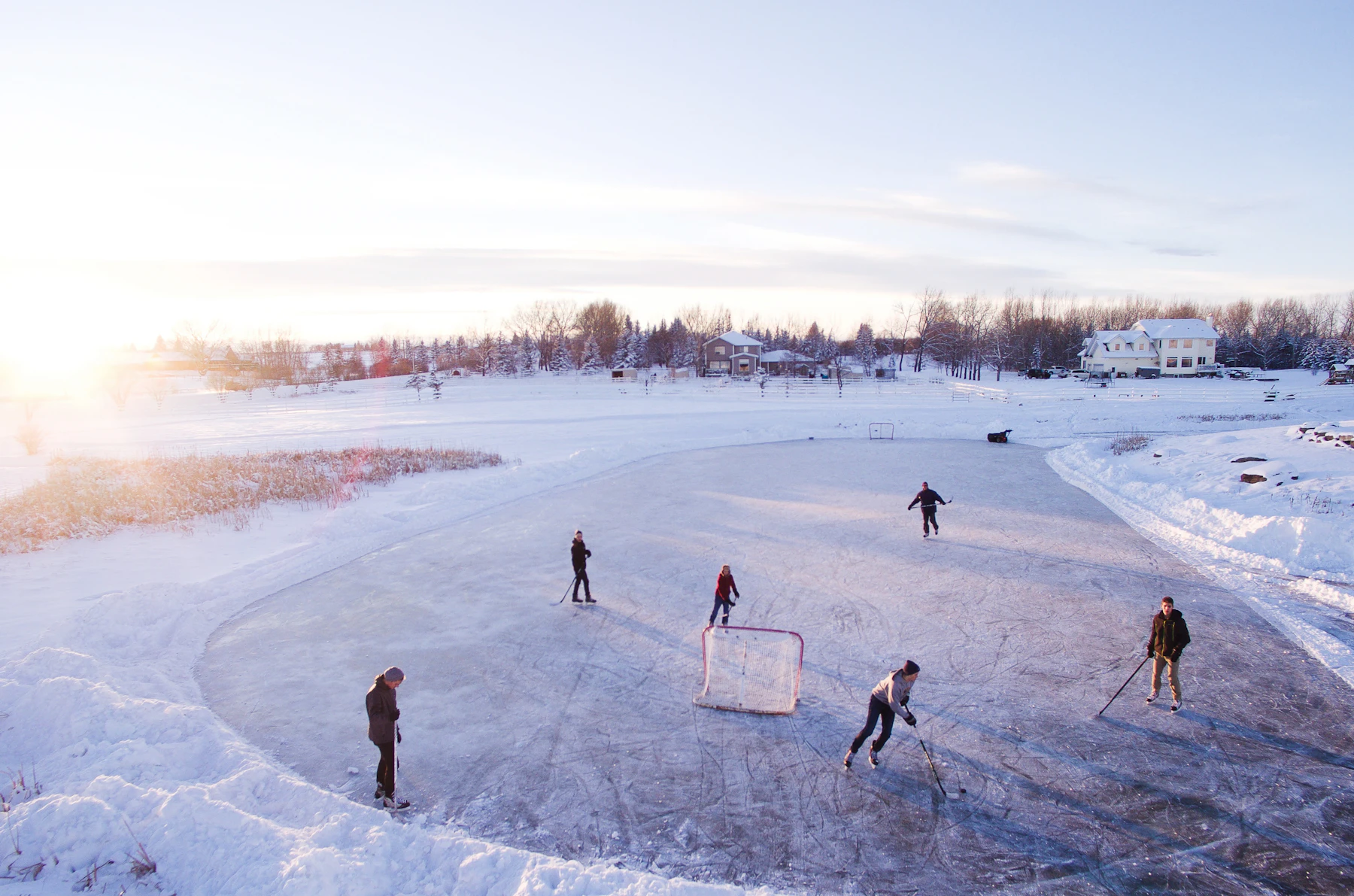 Skaters around a hockey net on a shoveled patch of outdoor ice at sunrise, with snowbanks and a house behind