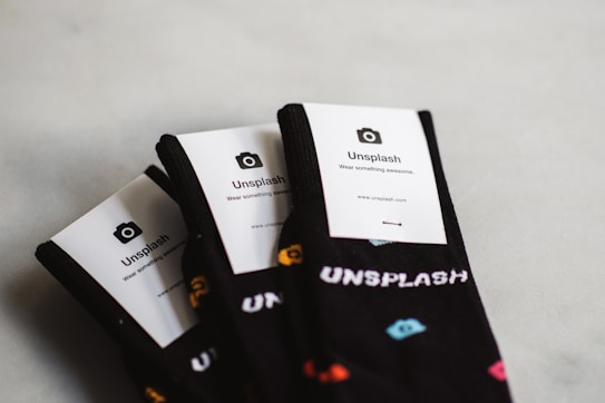Three pairs of black socks are neatly arranged, each with a white card label that has the word 'Unsplash' printed on it, along with a camera icon and some text below. The socks have small, colorful shapes printed on them.