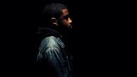 A person in a dark environment wearing a denim jacket and a hooded sweatshirt. The person is facing to the right with only the side profile visible, and the lighting highlights only parts of their face and upper body while the rest of the image is engulfed in darkness.