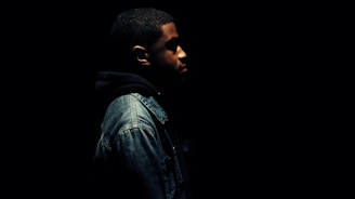A person in a dark environment wearing a denim jacket and a hooded sweatshirt. The person is facing to the right with only the side profile visible, and the lighting highlights only parts of their face and upper body while the rest of the image is engulfed in darkness.