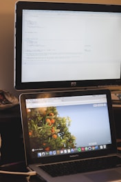 A workspace setup with two screens. The top monitor displays multiple lines of computer code in an editor. The bottom laptop screen shows a desktop with an open browser tab displaying an image of an orange tree with fruits against a clear blue sky. The focus is on dual-tasking between coding and viewing the webpage.